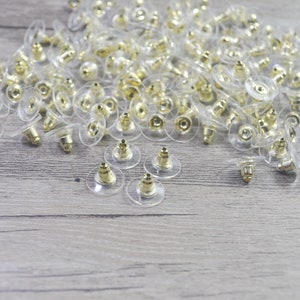 May include: A large quantity of clear plastic earring backs with gold-colored metal posts. The backs are scattered on a light brown wooden surface.