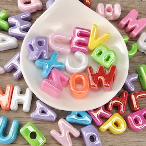 Puede incluir: Una colección de cuentas de alfabeto brillantes y coloridas en varios colores, incluyendo rosa, verde, azul y naranja. Algunas cuentas están dentro de un plato blanco en forma de lágrima, mientras que otras están esparcidas sobre una superficie de madera. Las cuentas tienen forma de letras.