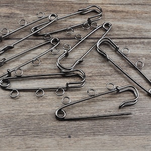 May include: A set of ten black safety pins with multiple loops on each pin. The pins are arranged in a random pattern on a wooden surface.
