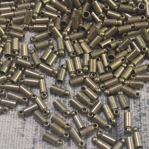 Puede incluir: Una gran cantidad de pequeños tapones metálicos de color dorado antiguo con una bola redonda en un extremo. Los tapones están esparcidos sobre una superficie de tela texturizada.