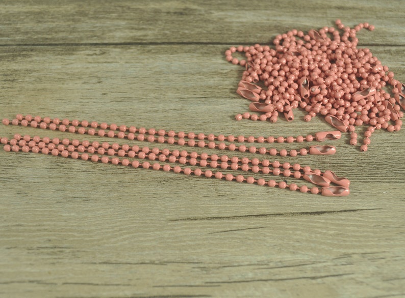 May include: A pile of pink metal chain with a few strands laid out on a wooden surface. The chain is made of small, round links.