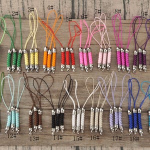 May include: A collection of 19 colorful braided phone charms with silver-toned metal accents. Each charm features a different color cord, including green, orange, red, pink, purple, blue, and brown. The charms are arranged on a wooden surface.