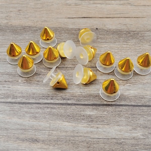 Könnte beinhalten: Eine Sammlung goldfarbener, pyramidenförmiger Metallnieten mit durchsichtigen Kunststoffsockeln. Die Nieten sind auf einer Holzoberfläche angeordnet.