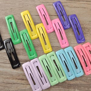 Könnte beinhalten: Eine Sammlung farbenfroher Haarspangen in verschiedenen Farbtönen, darunter Schwarz, Grün, Gelb, Rosa, Lila und Blau. Die Spangen sind rechteckig mit einem mittigen Ausschnitt, angeordnet auf einer Holzoberfläche.