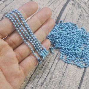 May include: A close-up of a hand holding a light blue metal chain. The chain is made of small, round beads and has a silver finish. There is a pile of the same chain in the background.