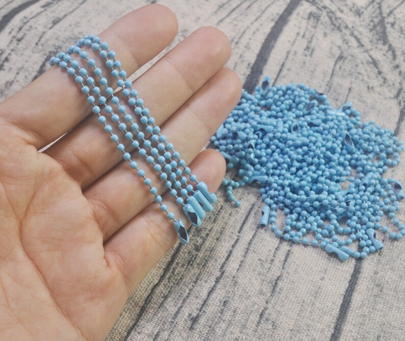 May include: A close-up of a hand holding a light blue metal chain. The chain is made of small, round beads and has a silver finish. There is a pile of the same chain in the background.