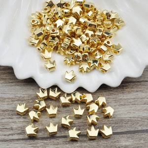 May include: A pile of gold crown-shaped beads on a white surface. A smaller group of the same beads are scattered on a wooden surface.