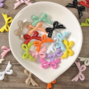 May include: A selection of colourful bow-shaped embellishments in shades of pink, yellow, and green, are arranged on a white plate and a wooden surface. Some bows have pins attached. These decorative bows are suitable for crafts and accessories.