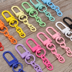 May include: A collection of colorful metal swivel clasps with matching chain links. The clasps are in various colors, including red, orange, yellow, green, blue, pink, purple, white, and black. The clasps are arranged on a wooden surface.
