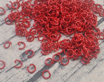 Könnte beinhalten: Eine Ansammlung kleiner, leuchtend roter Sprungringe, die auf einer strukturierten, hellgrauen Oberfläche verstreut sind. Die Ringe sind offen und bereit für die Verwendung in der Schmuckherstellung oder anderen Bastelarbeiten. Die Nahaufnahme hebt die leuchtende Farbe und den metallischen Glanz hervor.