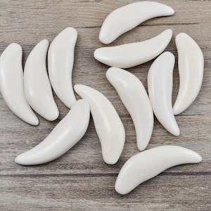 May include: Eleven smooth, white, crescent-shaped objects are arranged on a light brown wooden surface. Each piece has a curved, banana-like form. The objects are similar in shape.