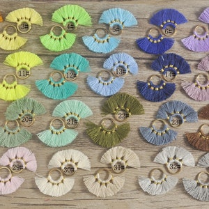 Może przedstawiać: Zestaw 34 kolorowych kolczyków z frędzlami i złotymi obręczami. Każdy kolczyk ma inny kolor i wzór frędzli.