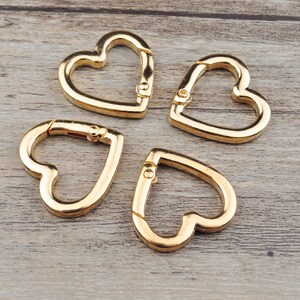 May include: Four gold-colored heart-shaped carabiner clips. Each clip has a spring-loaded gate for easy attachment. The clips are made of metal and are displayed on a light brown wooden surface.