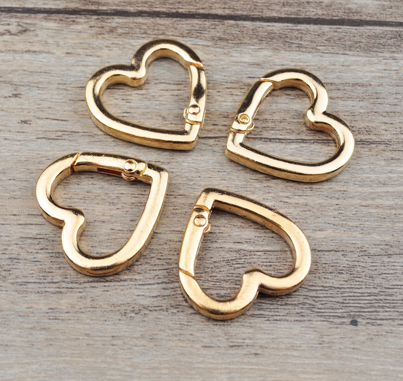 May include: Four gold-colored heart-shaped carabiner clips. Each clip has a spring-loaded gate for easy attachment. The clips are made of metal and are displayed on a light brown wooden surface.
