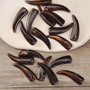 May include: A selection of dark brown, claw-shaped beads with a polished surface. Each bead features a small hole for threading. The beads have a natural, organic look, with variations in colour and pattern.