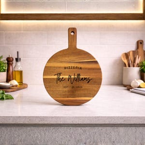 May include: A round wooden pizza board with a handle. The board features the text "Pizzeria The Williams EST. 2024" in a stylish font. The board is made of light brown wood and is displayed on a kitchen countertop.