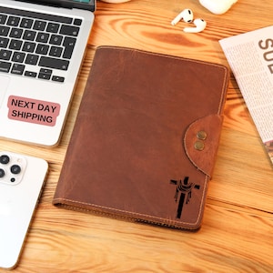 May include: A brown leather journal with a cross design embossed on the front cover. The journal has a flap closure with two metal snaps.