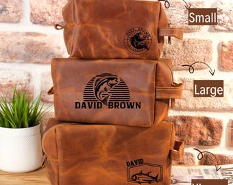 Peut inclure: Trois trousses de toilette en cuir marron de différentes tailles empilées. La trousse supérieure, étiquetée "Small", porte un logo circulaire avec le texte "ALEX BROWN". La trousse du milieu, étiquetée "Large", porte un logo de poisson et le texte "DAVID BROWN". La trousse inférieure, étiquetée "XLarge", porte un logo de poisson et le texte "DAVID".