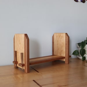 May include: A wooden bookend with a natural finish. The bookend is made of light and dark wood, with a rectangular base and two upright supports. The supports have a unique angled design. The bookend is approximately 10 inches wide and 6 inches tall.