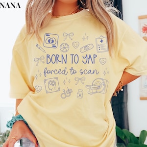 May include: A light yellow t-shirt with blue medical-themed graphics and the text "BORN TO YAP forced to scan". The design includes a pen, bows, hearts, and medical equipment illustrations.
