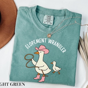 May include: Light green t-shirt with the text "ELOPEMENT WRANGLER" and a cartoon duck wearing a pink cowboy hat and boots, holding a lasso. A smaller duck is next to it. A gold necklace is on the shirt.