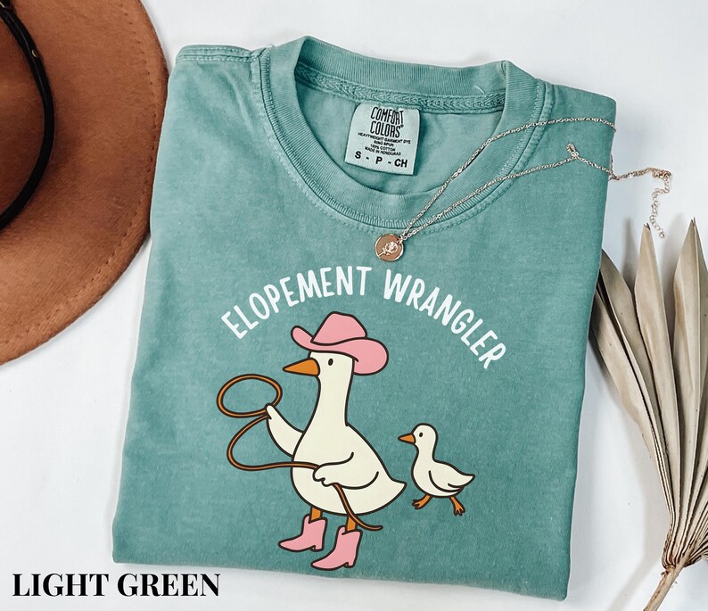 Puede incluir: Camiseta verde claro con el texto "ELOPEMENT WRANGLER" y un pato de dibujos animados con sombrero y botas de vaquero rosas, sosteniendo un lazo. Hay un patito m&aacute;s peque&ntilde;o al lado. Un collar dorado est&aacute; en la camisa.