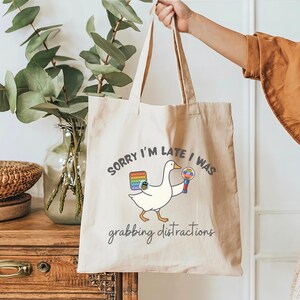 May include: Beige canvas tote bag with a cartoon goose holding a pop-it toy and a rainbow lollipop. Text on the bag reads "SORRY I'M LATE I WAS grabbing distractions". The bag has two long handles.