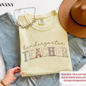 May include: A pale yellow t-shirt featuring the text kindergarten TEACHER in a faux patchwork design with floral and plaid patterns. The shirt is displayed with blue denim jeans, a brown fedora hat, and a silver-toned floral necklace.