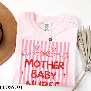 May include: A light pink t-shirt with a graphic that reads "MOTHER BABY NURSE" in red letters. The design includes a pink bow and vertical pink stripes. A gold necklace is draped across the shirt.