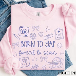 May include: Light pink sweatshirt with the text "BORN TO YAP forced to scan" in blue. The design includes medical-themed doodles such as a brain, a heart, and a CT scanner. The sweatshirt is made of a soft material.