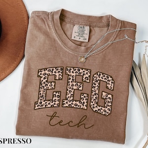 May include: Espresso-colored t-shirt with the text "EEG tech" in a leopard print design. The shirt has a crew neck and short sleeves. A silver necklace with a pendant is draped across the shirt.