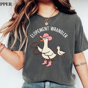 Puede incluir: Camiseta gris oscuro con un dibujo de un ganso de dibujos animados con sombrero y botas rosas, sosteniendo un lazo, con el texto "Elopement Wrangler". Un ganso m&aacute;s peque&ntilde;o camina al lado. La camiseta es de estilo casual.
