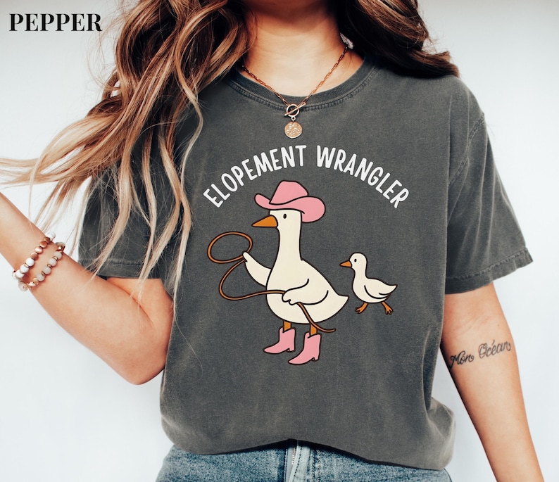 Puede incluir: Camiseta gris oscuro con un dibujo de un ganso de dibujos animados con sombrero y botas rosas, sosteniendo un lazo, con el texto "Elopement Wrangler". Un ganso m&aacute;s peque&ntilde;o camina al lado. La camiseta es de estilo casual.