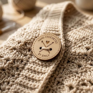 May include: A beige crocheted garment featuring a round wooden button with the engraved text "HAND CROCHETED BY EMILY" and a graphic of two crossed crochet hooks with a small heart in the centre, attached with a light-coloured thread.