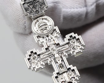 May include: Detailed silver Orthodox cross pendant. The cross features intricate religious iconography, including a central depiction of Jesus Christ on the cross, and other religious figures. The pendant is held by a white glove.