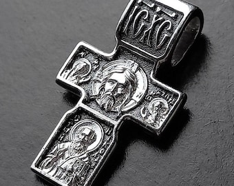 May include: A silver-colored pendant in the shape of a cross. The cross features detailed engravings of religious figures and text. The pendant has a loop at the top for a chain.