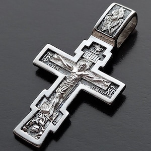 Puede incluir: Un colgante de plata detallado en forma de cruz, con una representación de Jesucristo. La cruz tiene grabados intrincados y una argolla para una cadena. El diseño incluye símbolos religiosos y texto.