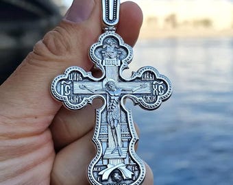 May include: A detailed silver pendant in the shape of a cross, featuring intricate religious iconography. The cross depicts a crucified figure and includes Cyrillic inscriptions. The pendant is held against a blurred background.