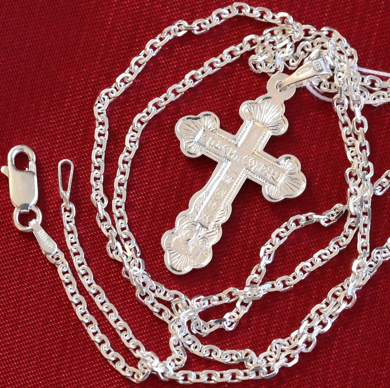 May include: A silver chain necklace with a silver cross pendant. The cross has intricate details and the inscription "Спаси и Сохрани" in Cyrillic script.