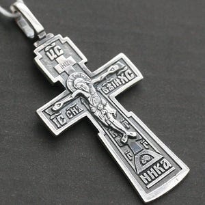 Op de afbeelding: Een orthodoxe kruishanger van zilverkleurig metaal met een reliëfvoorstelling van de kruisiging. Het ontwerp bevat ingewikkelde cyrillische letters en religieuze iconografie, met een oogje aan de bovenkant voor een ketting of koord.