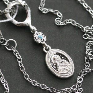 Könnte beinhalten: Eine silberfarbene Metallkette mit einer feinen Gliederkette und einem Anhänger. Der Anhänger besteht aus einer kleinen, ovalen Medaille mit einer religiösen Figur, die mit einem blütenförmigen Element mit hellblauem Stein verbunden ist.