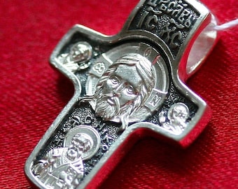 May include: A silver cross pendant with black enamel and detailed engravings. The cross features a central image of Jesus Christ and smaller images of saints around the edges. The inscription "Спаси Господи" is visible on the top of the cross.