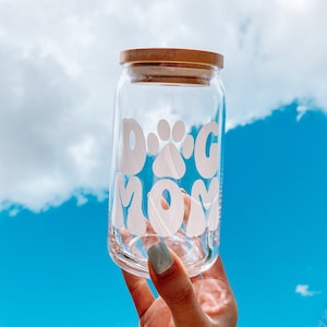 Pode incluir: Um frasco de vidro transparente com uma tampa de madeira e um adesivo de vinil branco que diz "Dog Mom" com uma pegada no meio.