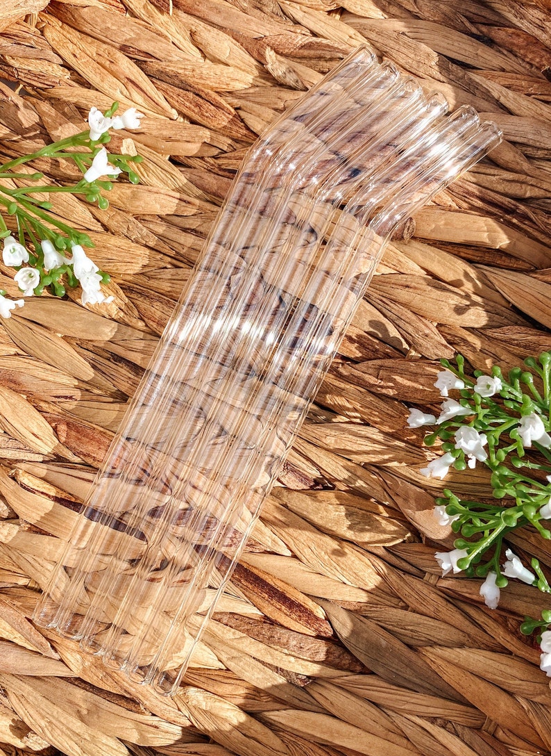 Pode incluir: Um conjunto de canudos de vidro transparente com uma curva na parte superior. Os canudos est&atilde;o dispostos em forma de leque sobre uma superf&iacute;cie de vime marrom.