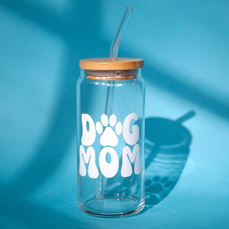 Pode incluir: Uma garrafa de &aacute;gua de vidro transparente com uma tampa de bambu e um design de impress&atilde;o de pata branca que diz "Dog Mom". Um canudo de vidro &eacute; inserido atrav&eacute;s da tampa.