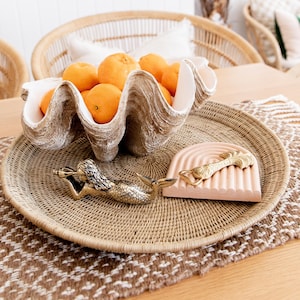May include: A decorative arrangement featuring a large, clam-shaped bowl filled with oranges. A woven tray holds a mermaid bottle opener and a pink, arched coaster with a gold accent. The scene is set on a wooden table.