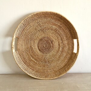 May include: A round, woven rattan tray with a natural, light brown color. The tray features a central circular design and two rectangular handles. The woven pattern creates a textured surface. This decorative tray is suitable for serving or display.