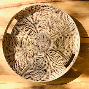 May include: A round, woven wicker tray with two built-in handles. The tray is light brown and has a circular pattern radiating from the center. The tray is sitting on a wooden surface.