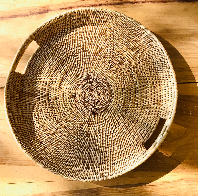 May include: A round, woven wicker tray with two built-in handles. The tray is light brown and has a circular pattern radiating from the center. The tray is sitting on a wooden surface.