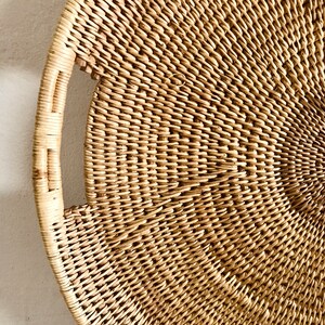 May include: A round, woven wicker tray with a handle. The tray is made of natural-colored wicker and has a tightly woven pattern. The handle is a curved shape, and the tray is likely used for serving or decoration.